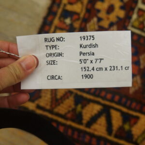 A hand holds a white tag over a geometric rug patterned in rust, blue, and gold colors.