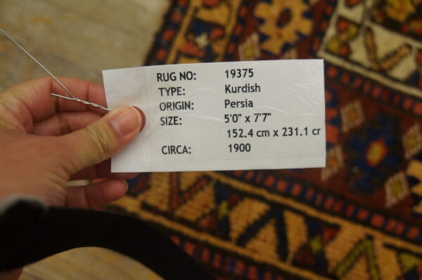 A hand holds a white tag over a geometric rug patterned in rust, blue, and gold colors.