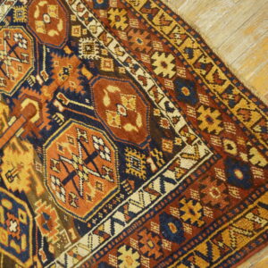 Geometric rug corner with complex borders in blue, rust, and gold sits on light wood planks.