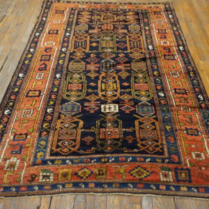 Low-pile tribal rug featuring bold navy, orange, and yellow geometric designs resting on rustic wood plank flooring.
