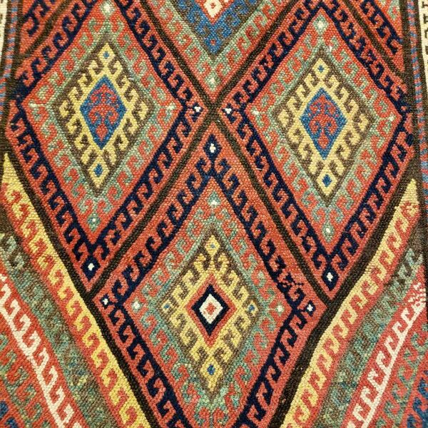 Geometric diamond rug pattern in vivid red, yellow, navy, and green with intricate hook motifs.