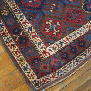 Red and blue geometric rug corner with cream borders resting on worn, light wood plank flooring.
