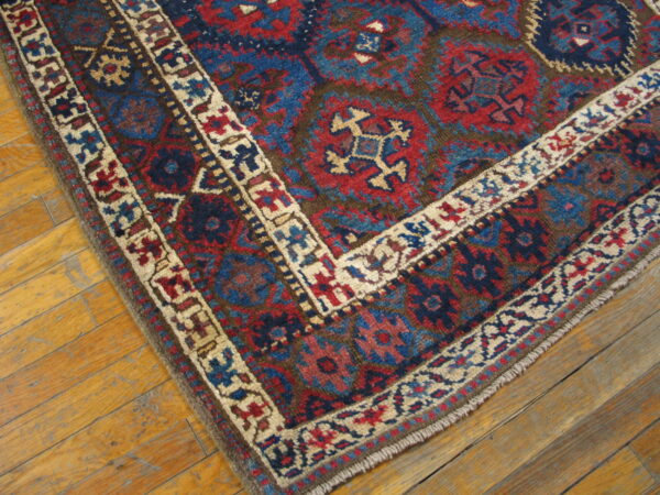 Red and blue geometric rug corner with cream borders resting on worn, light wood plank flooring.