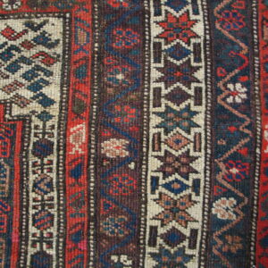 Tight weave close-up showing multiple geometric borders in red, blue, and off-white next to hardwood floor.