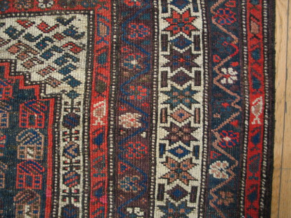 Tight weave close-up showing multiple geometric borders in red, blue, and off-white next to hardwood floor.