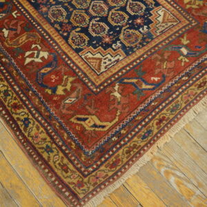 A close-up of a rustic red and navy rug featuring geometric medallions and stylized motifs on distressed wood floors.