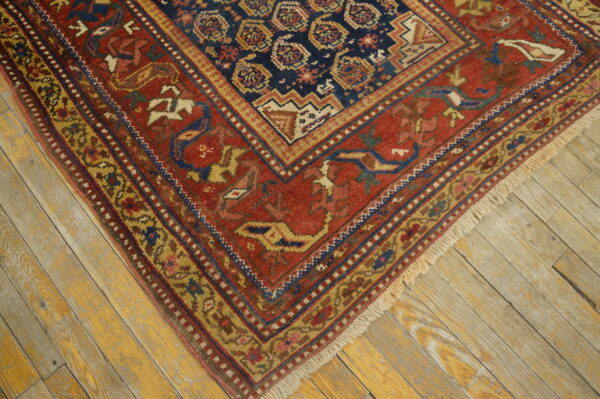 A close-up of a rustic red and navy rug featuring geometric medallions and stylized motifs on distressed wood floors.