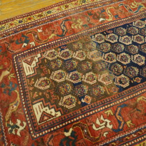 Geometric rug in shades of red, navy, brown, and gold displayed on yellow wood flooring.