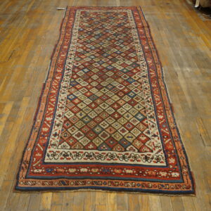 Red runner rug with multicolored geometric diamond field pattern rests on worn wood plank flooring.