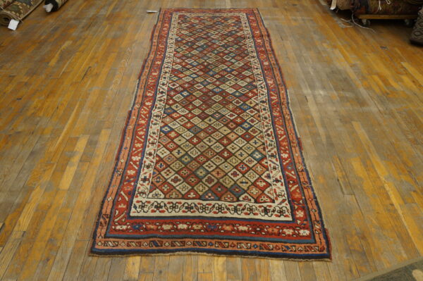 Red runner rug with multicolored geometric diamond field pattern rests on worn wood plank flooring.
