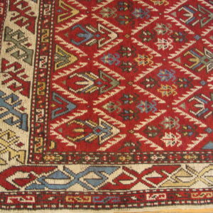 Detailed corner of a red geometric rug with diamond field pattern and multicolored borders resting on wood planks.