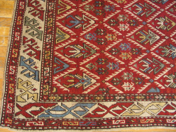 Detailed corner of a red geometric rug with diamond field pattern and multicolored borders resting on wood planks.
