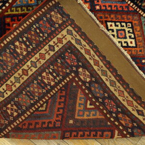 A multi-colored traditional geometric rug corner is folded, revealing a brown canvas backing over wood flooring.