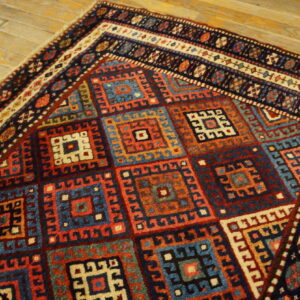 Geometric rug with nested square designs in blue, red, orange, and cream on rustic wood plank flooring.