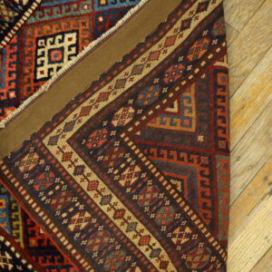 Complex geometric rug in deep reds, blues, and tan is partially folded on light wood flooring.
