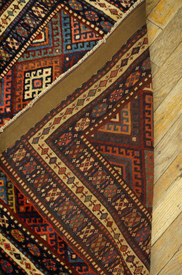 Complex geometric rug in deep reds, blues, and tan is partially folded on light wood flooring.