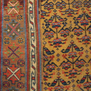 Detailed close-up of a low-pile rug in mustard yellow, red, dark blue, and tan geometric patterns next to wood.