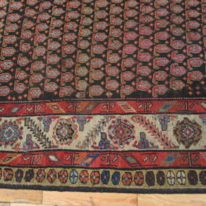 Dark rug featuring red repeating stylized motifs and detailed multi-colored borders on wood flooring.