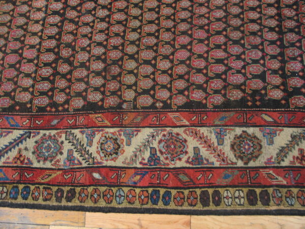 Dark rug featuring red repeating stylized motifs and detailed multi-colored borders on wood flooring.