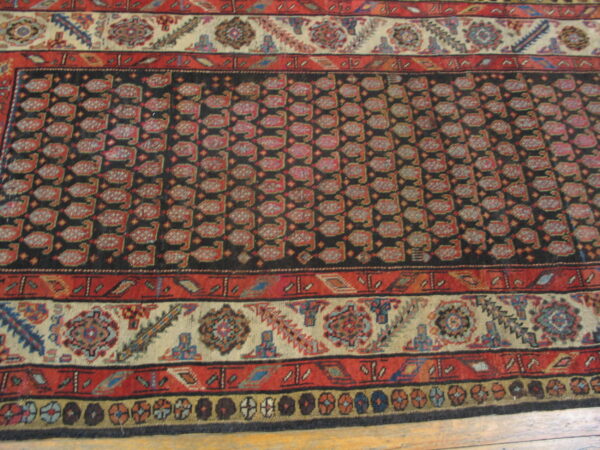 Traditional rug featuring a dark central panel with repeating red motifs, detailed cream and red borders, and wood flooring.