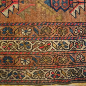 Muted, knotted rug edge with a rust field and complex floral borders in blue, red, tan, and brown.