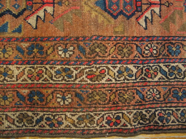 Muted, knotted rug edge with a rust field and complex floral borders in blue, red, tan, and brown.