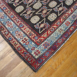 Dark rug with dense geometric patterns in red, blue, and cream sits on light brown wooden floorboards.