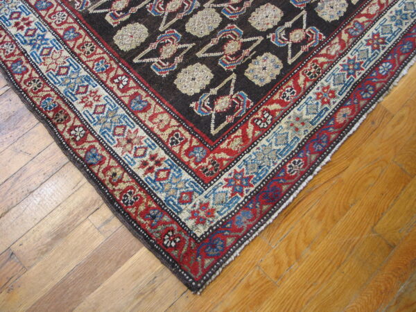 Dark rug with dense geometric patterns in red, blue, and cream sits on light brown wooden floorboards.