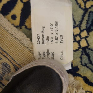 Ornate navy, gold, and teal rug close-up showing fringe, the checkered backing, a paper tag, and a dark shoe.