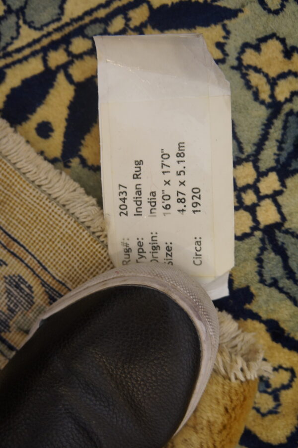 Ornate navy, gold, and teal rug close-up showing fringe, the checkered backing, a paper tag, and a dark shoe.