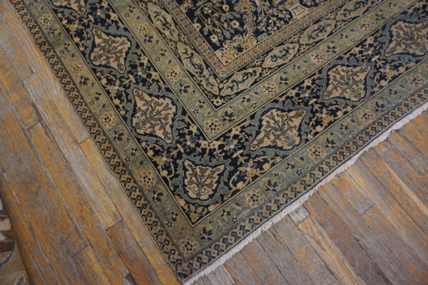 Dark blue and beige rug with intricate floral patterns and green borders rests on distressed wood flooring.