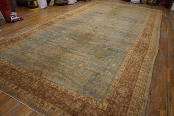 A low-pile traditional rug with a faded blue floral field and wide rust borders rests on wood planks.