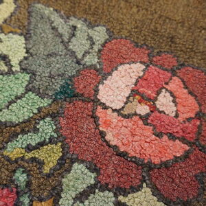 Close-up of a hooked rug showing a floral pattern in red, pink, and sage green on a dark brown background.