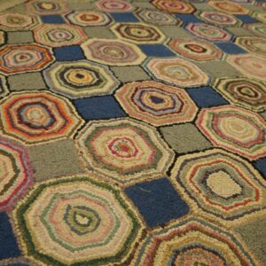 A multicolor hooked rug featuring geometric hexagons with concentric rings and contrasting blue and gray squares on wood flooring.