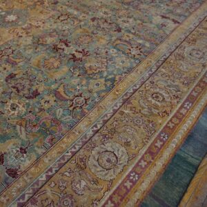 Faded gold, teal, and purple floral rug over a dark teal rug on peeling yellow wood planks.