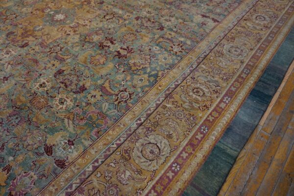 Faded gold, teal, and purple floral rug over a dark teal rug on peeling yellow wood planks.