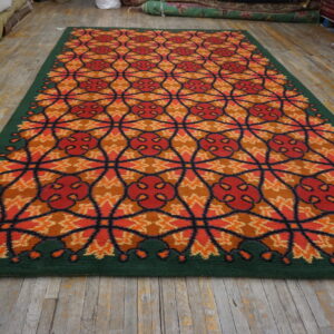 A colorful rug with an orange, red, and brown geometric pattern and a solid green border lies on a wood floor.