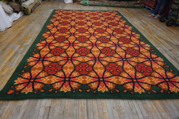 A colorful rug with an orange, red, and brown geometric pattern and a solid green border lies on a wood floor.