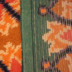 A high-pile orange geometric rug edge connects to a green binding and a multicolored flat-weave pattern.