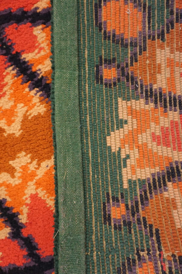 A high-pile orange geometric rug edge connects to a green binding and a multicolored flat-weave pattern.