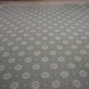Light sage rug with repeating white daisies and a detailed floral border on wood flooring.