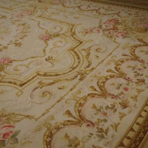 A large low-pile rug featuring a cream background with gold scrollwork and pink floral bouquets on wood flooring.