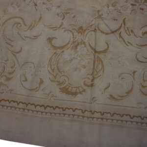 Muted beige rug with detailed tan baroque scrolls, floral patterns, and a dark scalloped border.