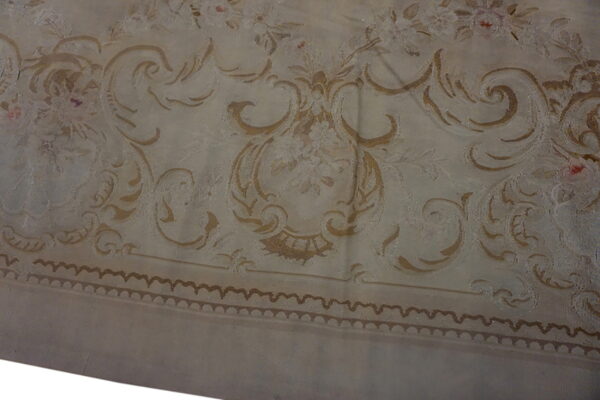 Muted beige rug with detailed tan baroque scrolls, floral patterns, and a dark scalloped border.