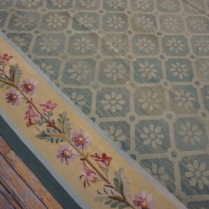 Low-pile blue lattice rug with a beige border displaying pink and purple floral needlework on wood flooring.