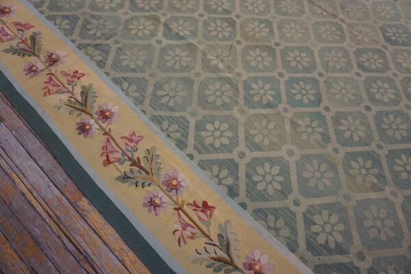 Low-pile blue lattice rug with a beige border displaying pink and purple floral needlework on wood flooring.