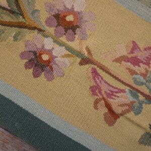 Flatweave rug detailed with purple and pink flowers on a yellow field and green striped border resting on wood floor.