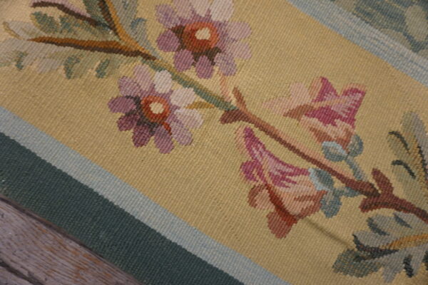 Flatweave rug detailed with purple and pink flowers on a yellow field and green striped border resting on wood floor.