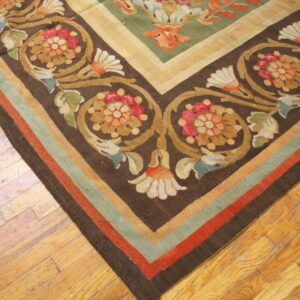 Flat-woven rug corner featuring dark brown, green, red, and gold scrolling floral patterns on wood flooring.