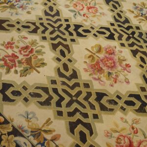 A cream rug displays dark brown interlocking geometric bands framing multicolored floral bouquets on a wood floor.